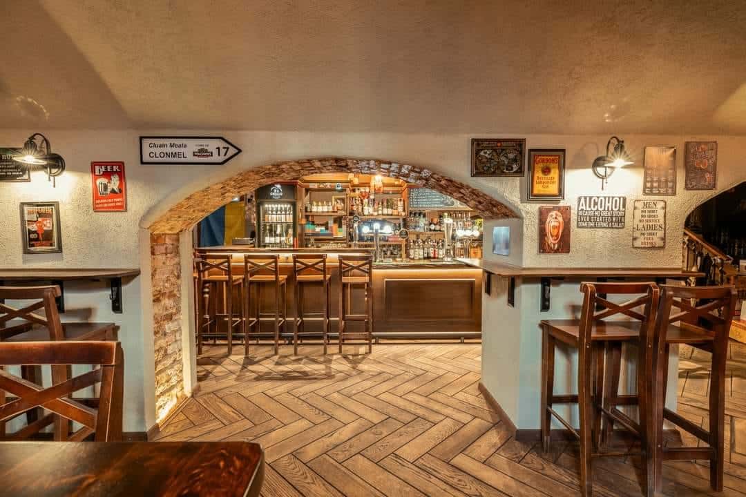 Rūsio baras su medine baro sritimi, aukštomis kėdėmis, arkinėmis plytų detalėmis ir plakatais ant sienų