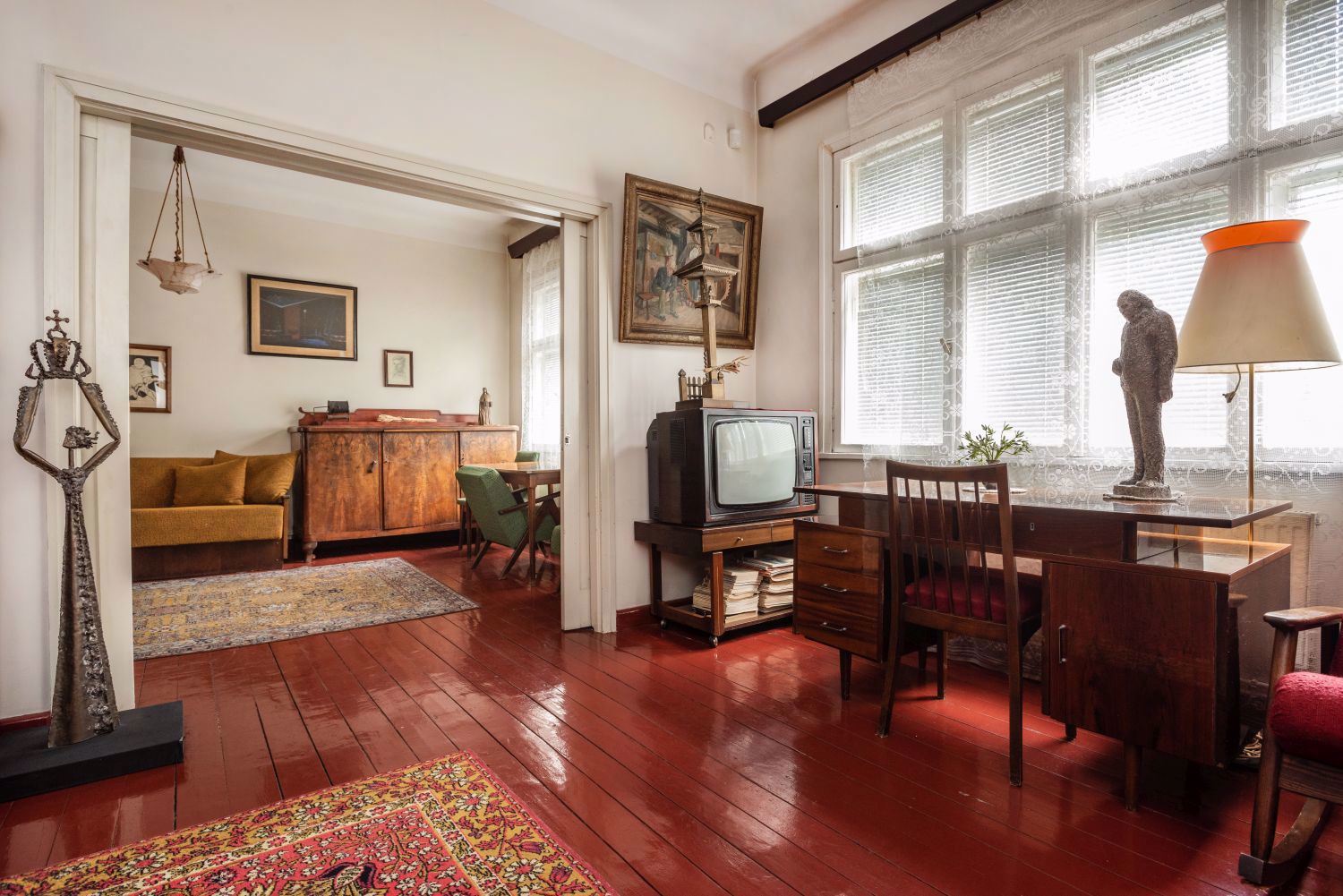 Juozo Grušo namų-muziejaus interjeras – erdvus kambarys su raudonmedžio spalvos grindimis, rašomuoju stalu prie lango, senovine televizija, mediniais baldais, kilimais ir meno kūriniais. Fone matomas svetainės kambarys su sofa ir spinta, išlaikantis autentišką XX a. vidurio namų atmosferą.